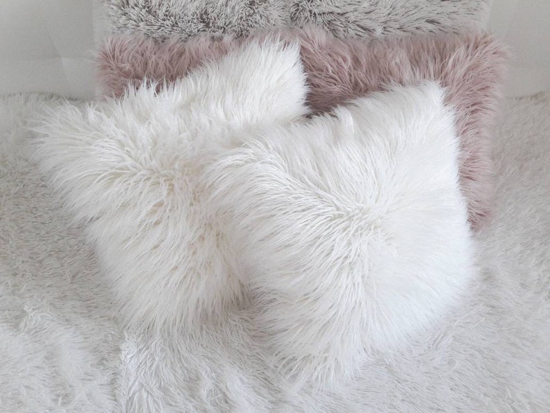 May include: Three fluffy throw pillows, one white and two in a light pink color, are arranged on a white fluffy surface. The pillows are made of a soft, plush material.