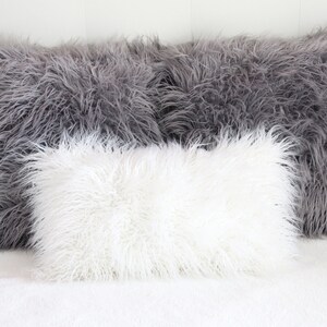 May include: Three fluffy pillows, one white and two gray, arranged on a white bedspread. The pillows are made of a soft, plush material that resembles fur.