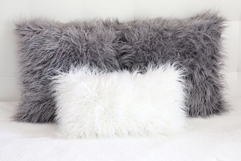 May include: Three fluffy pillows, one white and two gray, arranged on a white bedspread. The pillows are made of a soft, plush material that resembles fur.