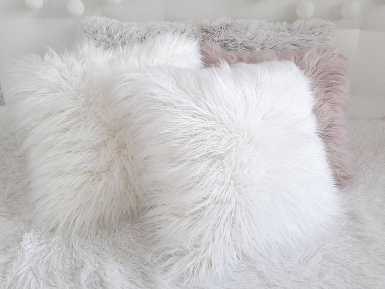 May include: Three fluffy throw pillows, one white, one light gray, and one light pink. The pillows are arranged on a white fluffy surface.
