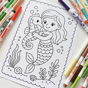 Op de afbeelding: Een kleurplaat met een zeemeermin die een zeepaardje omhelst, omringd door zeewier en bellen. Verschillende gekleurde stiften, waaronder rood, groen, oranje en blauw, liggen verspreid rond de pagina. De afbeelding staat op een wit oppervlak.