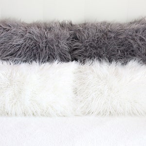 May include: Four fluffy pillows, two white and two gray, arranged on a white bedspread. The pillows are made of a soft, plush material and have a long, shaggy texture.