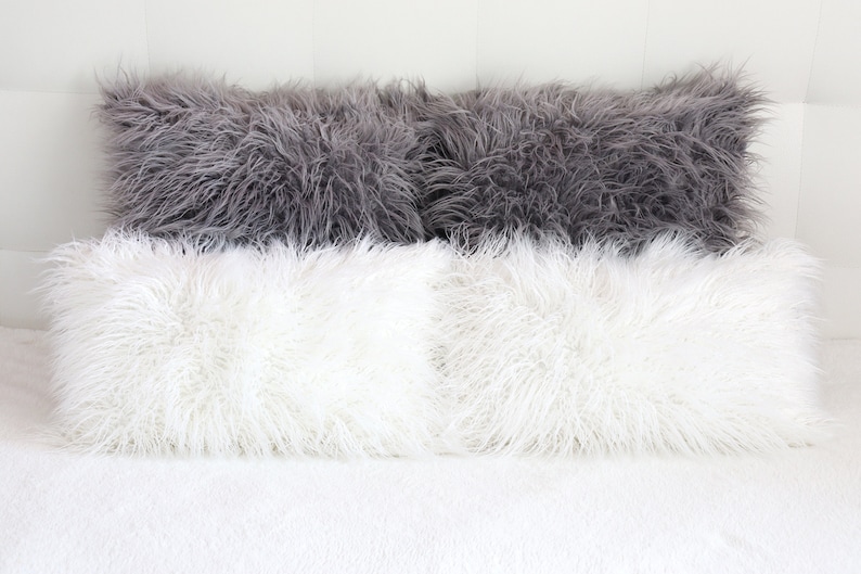 May include: Four fluffy pillows, two white and two gray, arranged on a white bedspread. The pillows are made of a soft, plush material and have a long, shaggy texture.