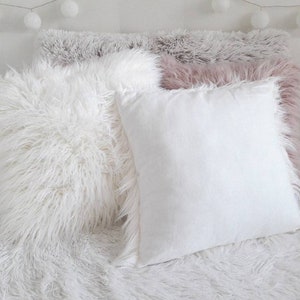 May include: Three decorative throw pillows with a fluffy, white faux fur texture. The pillows are arranged on a white faux fur surface.