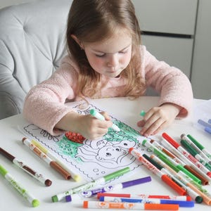 Op de afbeelding: Een kind kleurt een zeemeermin illustratie met een verscheidenheid aan kleurrijke stiften op een witte tafel. De stiften liggen in een doorzichtige plastic bak en verspreid rond de kleurplaat.