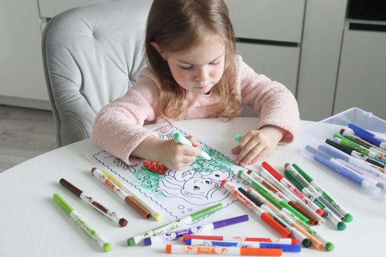 Op de afbeelding: Een kind kleurt een zeemeermin illustratie met een verscheidenheid aan kleurrijke stiften op een witte tafel. De stiften liggen in een doorzichtige plastic bak en verspreid rond de kleurplaat.