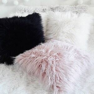 May include: Three fluffy throw pillows in black, white, and pink. The pillows are arranged on a white fluffy bedspread.
