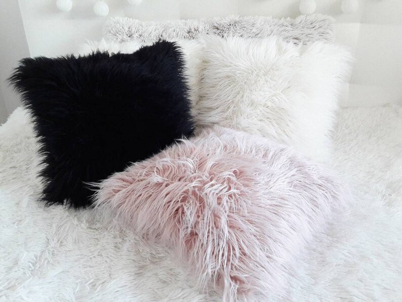 May include: Three fluffy throw pillows in black, white, and pink. The pillows are arranged on a white fluffy bedspread.