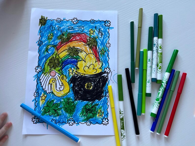 May include: A coloring page featuring a leprechaun, a pot of gold, and a rainbow. The page is surrounded by green shamrocks and stars. There are several green, blue, red, and black markers on a white surface.