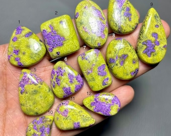 May include: A collection of twelve polished gemstones in various shapes, including teardrop, oval, and rectangular. The stones feature a vibrant yellow-green base color with striking purple veining and patterns. The gemstones are held in a hand.