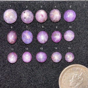 Może przedstawiać: Piętnaście różowych kamieni szlachetnych z rubinu gwiazdowego o różnych rozmiarach jest wyświetlanych na ciemnej powierzchni. Kamienie szlachetne są okrągłe lub owalne, z efektem gwiazdy widocznym na ich powierzchniach. Moneta jest dołączona do porównania rozmiarów.