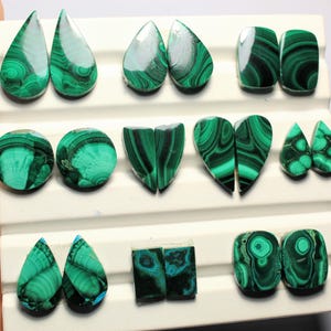 May include: Assortment of polished malachite gemstones in various shapes, including teardrops, ovals, and hearts. The stones display a vibrant green color with distinctive swirling patterns. The gemstones are arranged on a white display surface.