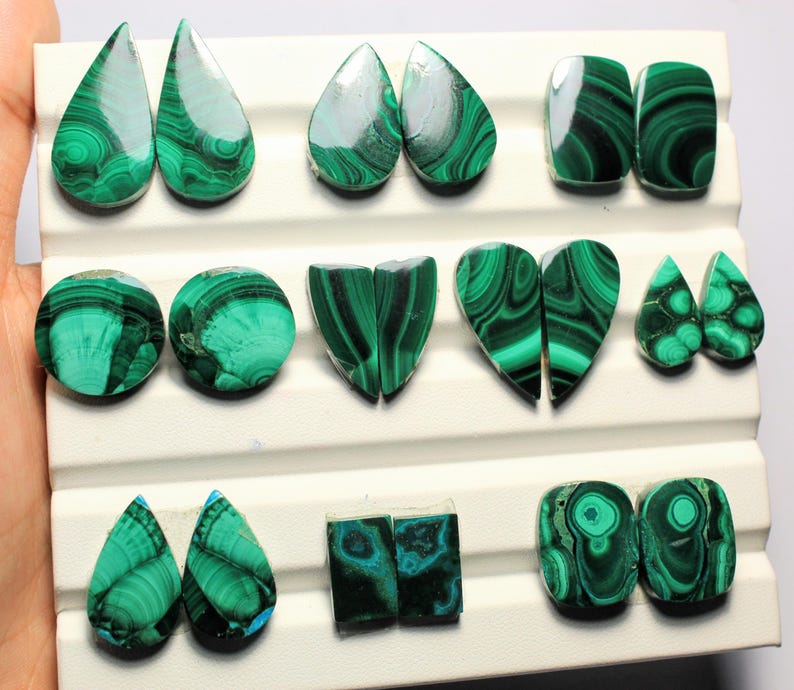 May include: Assortment of polished malachite gemstones in various shapes, including teardrops, ovals, and hearts. The stones display a vibrant green color with distinctive swirling patterns. The gemstones are arranged on a white display surface.