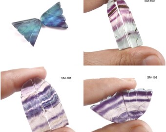 May include: A collection of carved fluorite gemstone pieces. The pieces include a butterfly, and several leaf-shaped carvings. The gemstones display shades of purple, blue, and green. Each piece is held by a person's fingers.