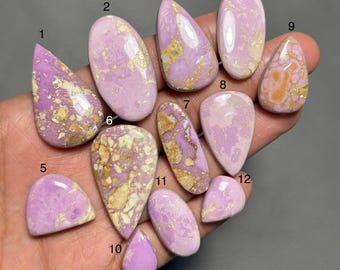 May include: A collection of twelve polished gemstones in various shapes, including oval, teardrop, and irregular forms. The stones display a soft lavender hue with golden-brown matrix patterns. The gemstones are held in a hand.
