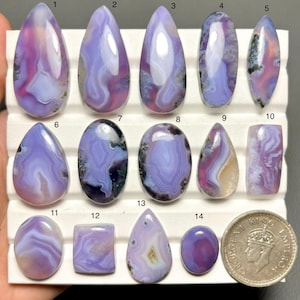 May include: A collection of fourteen polished agate gemstones in various shapes, including teardrop, oval, and rectangular. The stones display a range of colors, primarily lavender and white, with some featuring darker inclusions. A coin is included for size reference.