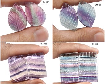 May include: Four pairs of carved fluorite gemstones in leaf and rectangular shapes. The stones display a range of colors, including purple, pink, and blue, with intricate leaf vein details. Each pair is held by a hand, with labels SM-107, SM-108, SM-109, and SM-110.
