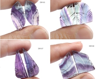 May include: Four pairs of carved fluorite gemstones in various shapes. The stones are held by fingers and display shades of purple, white, and blue. Each pair is labeled with a white text: SM-95, SM-96, SM-97, and SM-98.