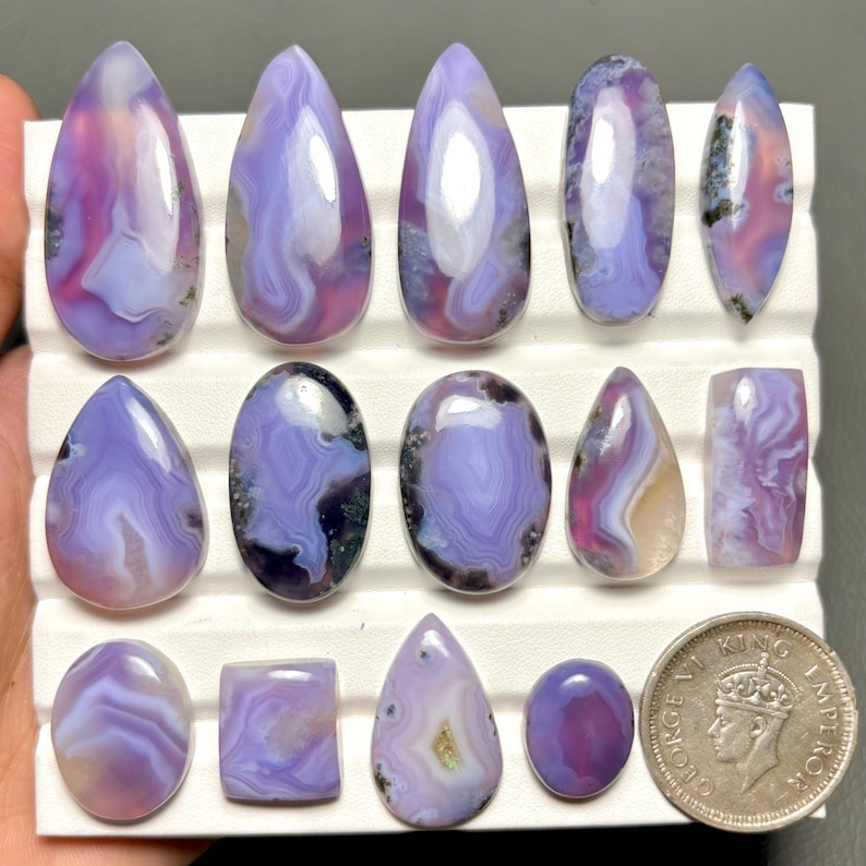 May include: A collection of polished gemstones in various shapes, including teardrop, oval, and rectangular. The stones display a mix of lavender, purple, and white colors with swirling patterns. A coin is included for size reference.