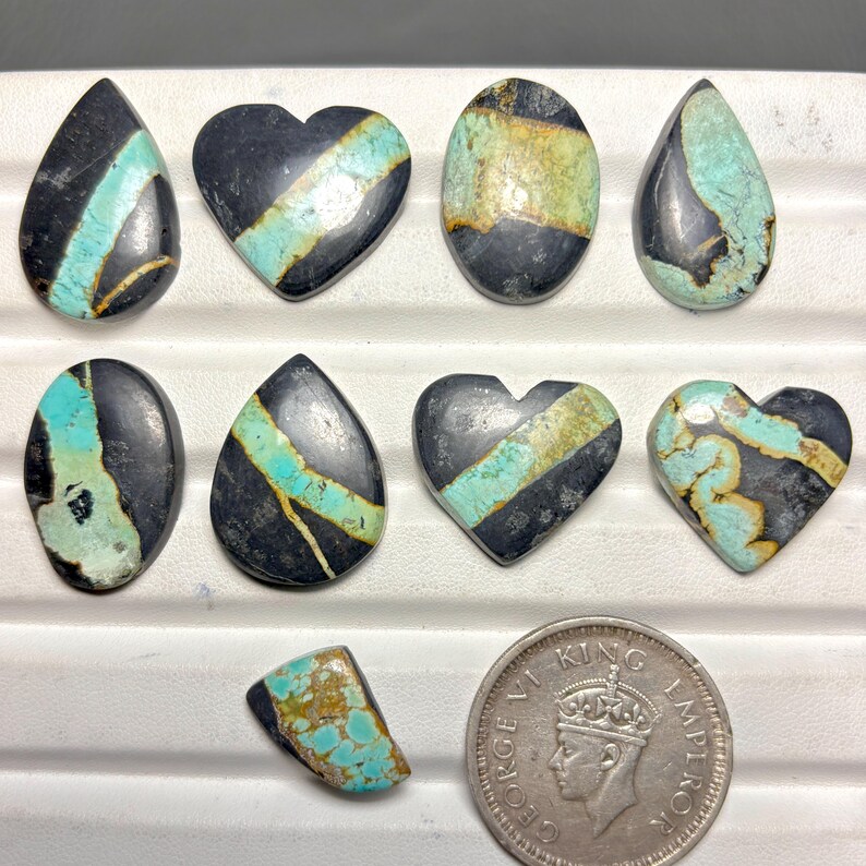 May include: A collection of polished gemstones in various shapes, including teardrop, oval, and heart. The stones feature a striking contrast of black and turquoise, with some displaying a band of turquoise across the center. A coin is included for size reference.