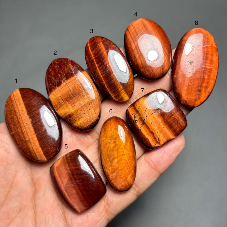 May include: A collection of eight polished tiger's eye gemstones in various shapes, including oval and rectangular. The stones display rich hues of brown, gold, and red, with visible chatoyancy. The gemstones are held in a hand.