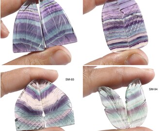 May include: Four pairs of polished fluorite gemstones in shades of purple, green, and white. Each pair is held by a hand, showcasing the natural patterns and carved details. The gemstones are labeled SM-91, SM-92, SM-93, and SM-94.