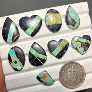 May include: A collection of polished gemstones in various shapes, including teardrop, oval, and heart. The stones feature a striking contrast of black, turquoise, and gold colors. A coin is included for size reference.