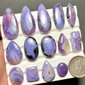 May include: A collection of polished agate gemstones in various shapes, including teardrop, oval, and rectangular. The stones display a range of colors, primarily lavender, purple, and white, with some featuring pink and brown hues. A coin is included for size reference.