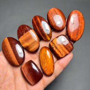 May include: A collection of polished tiger's eye gemstones in various shapes, including oval and rectangular. The stones display rich hues of brown, orange, and gold, with visible chatoyancy. The gemstones are held in a hand against a gray background.