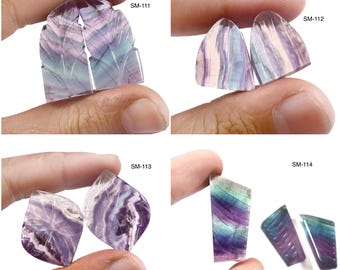 May include: Four pieces of polished fluorite gemstone, each held by a hand. The stones display a range of colors, including purple, green, and white, with visible patterns and textures. Each piece has a unique shape and cut, with the labels "SM-111", "SM-112", "SM-113", and "SM-114".