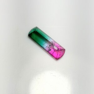 K&ouml;nnte beinhalten: Ein rechteckiger, mehrfarbiger Edelstein mit polierter, facettierter Oberfl&auml;che. Der Edelstein geht von einem tiefen Gr&uuml;n an einem Ende in ein leuchtendes Pink am anderen &uuml;ber, mit einem klaren, durchscheinenden Abschnitt dazwischen. Der Edelstein ist etwa 2,5 cm lang.