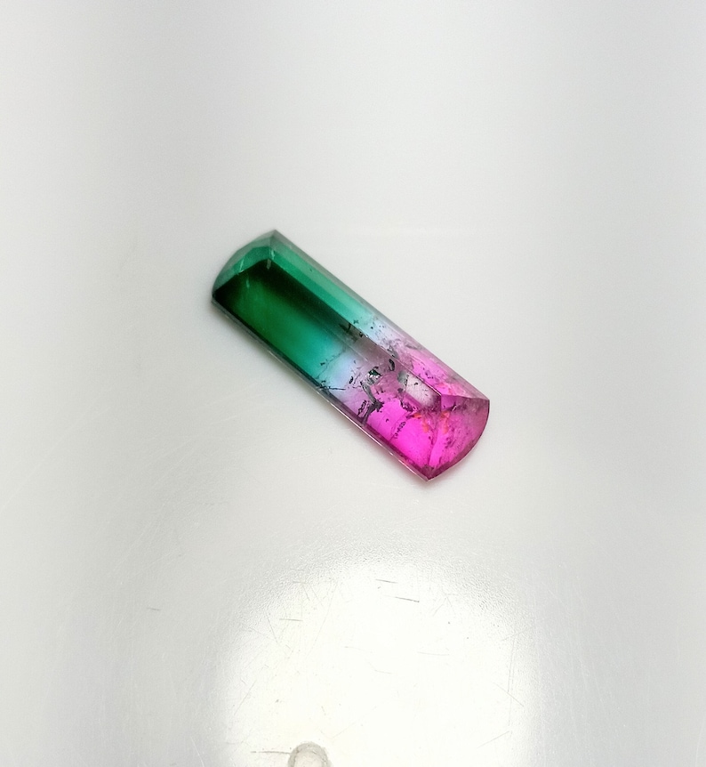 K&ouml;nnte beinhalten: Ein rechteckiger, mehrfarbiger Edelstein mit polierter, facettierter Oberfl&auml;che. Der Edelstein geht von einem tiefen Gr&uuml;n an einem Ende in ein leuchtendes Pink am anderen &uuml;ber, mit einem klaren, durchscheinenden Abschnitt dazwischen. Der Edelstein ist etwa 2,5 cm lang.