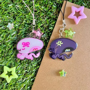 Peut inclure: Deux breloques de téléphone en acrylique représentant des créatures extraterrestres. L'une est rose clair avec un nœud, l'autre est violet foncé avec une expression en colère. Elles ont des fermoirs en métal argenté, des cordons et des perles en étoile.