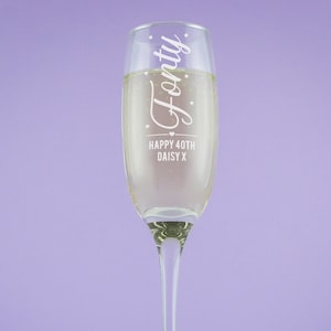 May include: A clear champagne flute filled with a sparkling drink. The glass is etched with the word "Forty" and the message "Happy 40th Daisy X". The flute has a long stem and sits against a lilac background.
