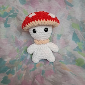 Puede incluir: Una figura de hongo hecha a mano con ganchillo, con un sombrero rojo con lunares blancos. El hongo tiene un cuerpo blanco, ojos negros y un cuello beige. El peluche está sobre un fondo floral colorido.