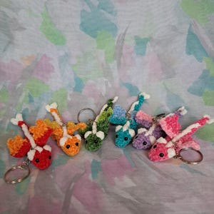 May include: A selection of crochet dragon keyrings in various colours. Each dragon has a unique colour scheme, including red, orange, green, blue, purple, and pink, with white accents. The keyrings are attached to silver metal rings.