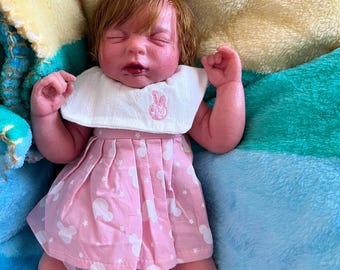 May include: A pink and white dress with a white bib and a pink bunny design. The dress is worn by a realistic baby doll with red hair and closed eyes. The doll is lying on a multi-colored blanket.
