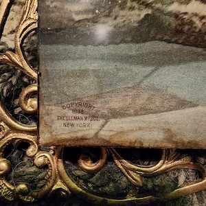 May include: An antique framed print with a gold-coloured ornate frame. The print features a landscape scene. The frame has a decorative, swirling design. The text "COPYRIGHT 1898 THE ULLMAN MFG CO. NEW YORK" is visible.