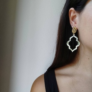 Puede incluir: Pendientes colgantes dorados con un diseño floral en la parte superior y una forma calada de cuatro hojas. Los pendientes se llevan en la oreja de una persona, con la correa negra de una camiseta sin mangas visible.