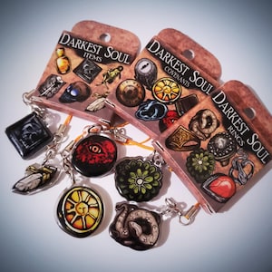 May include: A selection of Darkest Soul keychains and charms. The charms display diverse designs, such as an eye, a sun, a snake, and other game-related icons. The packaging reads "Darkest Soul Items", "Covenants", and "Rings".