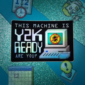 May include: A black sticker with the text "THIS MACHINE IS Y2K READY ARE YOU?" in holographic lettering. The sticker features a pixelated computer graphic with a screen displaying a warning symbol. Additional pixelated graphics of a clock, a building, and a lightbulb are also present.