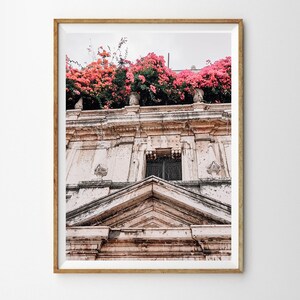 May include: A pink bougainvillea plant growing over a white building with a decorative archway and a window with a black metal gate. The building has a weathered, aged look.