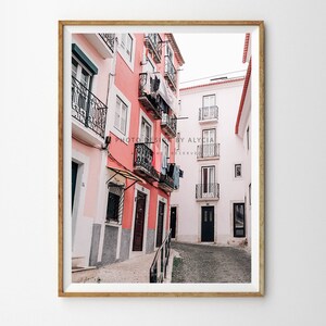 May include: A narrow, cobbled street in a European city with pink and white buildings. The buildings have balconies with black wrought iron railings and laundry hanging on lines.