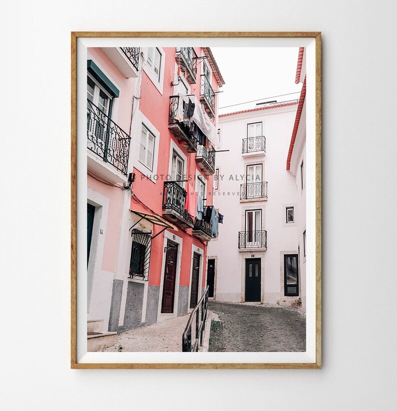 May include: A narrow, cobbled street in a European city with pink and white buildings. The buildings have balconies with black wrought iron railings and laundry hanging on lines.