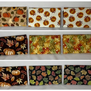May include: Nine fabric wallets with autumn-themed patterns. Designs include pumpkins, leaves, and berries in shades of orange, brown, and green. The wallets are rectangular and likely for holding cards or cash.