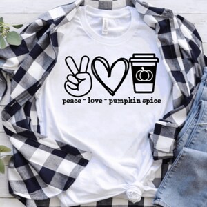 Könnte beinhalten: Weißes T-Shirt mit schwarzem Grafikdesign, das ein Friedenszeichen, ein Herz und eine Kaffeetasse mit einem Kürbis zeigt. Der Text lautet "peace - love - pumpkin spice". Das Shirt wird mit einem schwarz-weiß karierten Hemd und blauen Jeans kombiniert.
