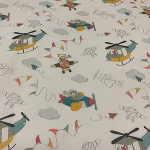 Op de afbeelding: Witte stof met een herhalend patroon van vliegtuigen en helikopters in verschillende kleuren, waaronder blauw, geel, rood en groen. De vliegtuigen en helikopters zijn zwart omlijnd en hebben een cartoonachtige stijl.
