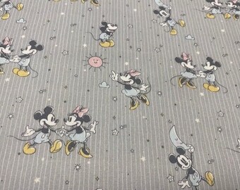 Op de afbeelding: Grijze stof met een herhalend patroon van Mickey Mouse en Minnie Mouse. De personages zijn in een cartoon-stijl en houden elkaars hand vast. De achtergrond is lichtgrijs met witte strepen en witte sterren.