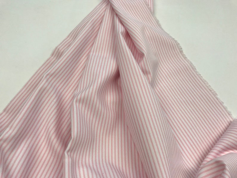 May include: Pink and white striped fabric with a subtle sheen. The fabric is folded and draped, showcasing the vertical stripes.