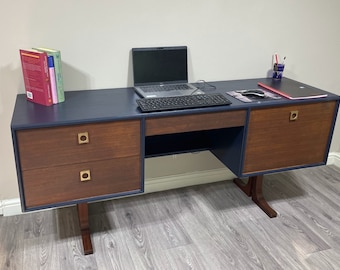 Può includere: Una scrivania blu scuro con un piano in legno e due cassetti sul lato sinistro. La scrivania ha un solo cassetto al centro e un armadio con un'unica porta sul lato destro. La scrivania ha ferramenta dorata e poggia su due gambe in legno su ogni lato. Un computer portatile, una tastiera e un mouse sono sulla scrivania.